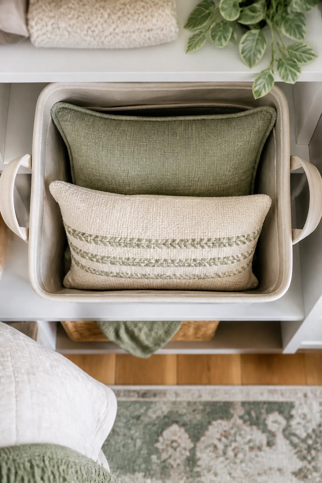 A fabric storage bin with handles on a closet shelf, two folded decorative pillows stacked inside it.