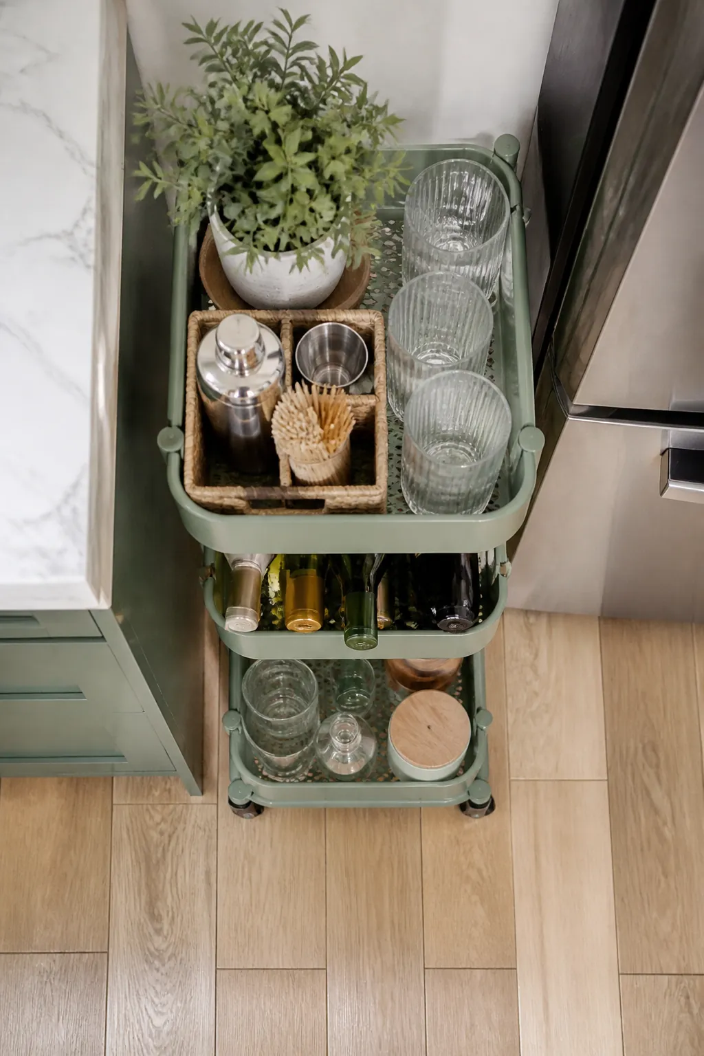 A narrow three-tier rolling cart beside a fridge holds glasses on top and wine bottles lying flat below.