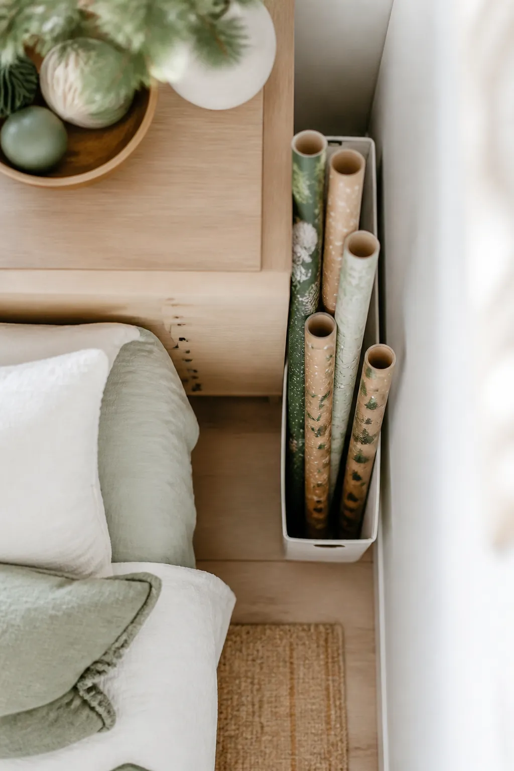 Several wrapping paper rolls standing upright in a narrow bin wedged into the gap beside a bedroom dresser.