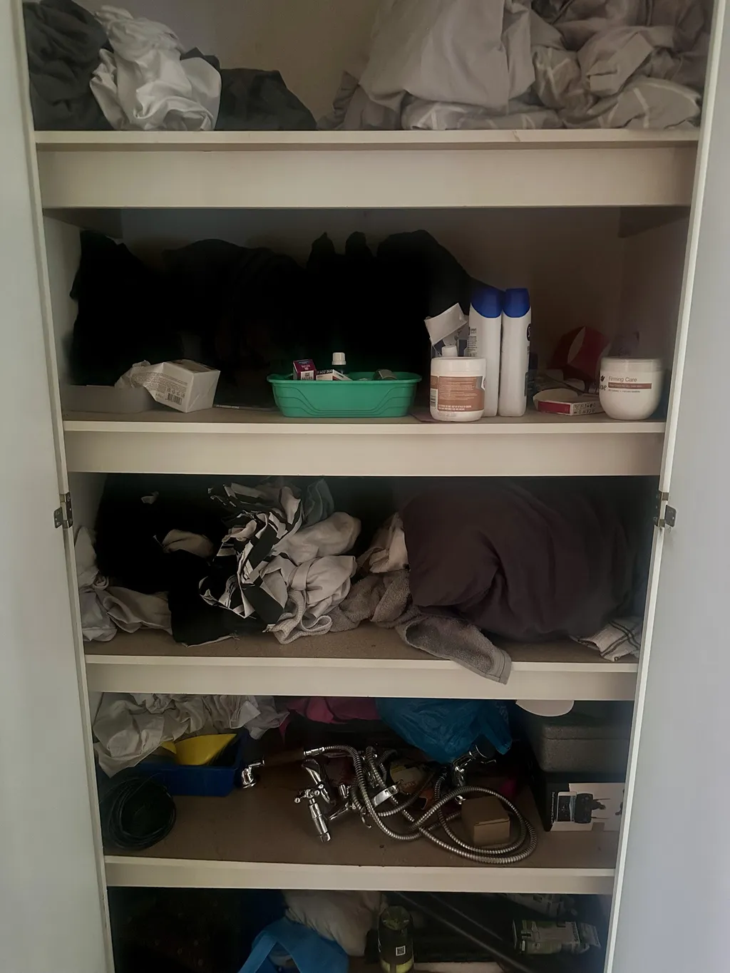 The author's linen closet as it is: bedding and towels piled unfolded on deep shelves, toiletries and a small green tray on one shelf, old replaced faucets and a shower hose on the bottom shelf.
